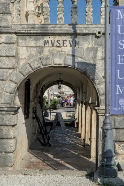 Ancient archway of the museum in Perast, Montenegro, with historic anchors in front and view of a busy street.