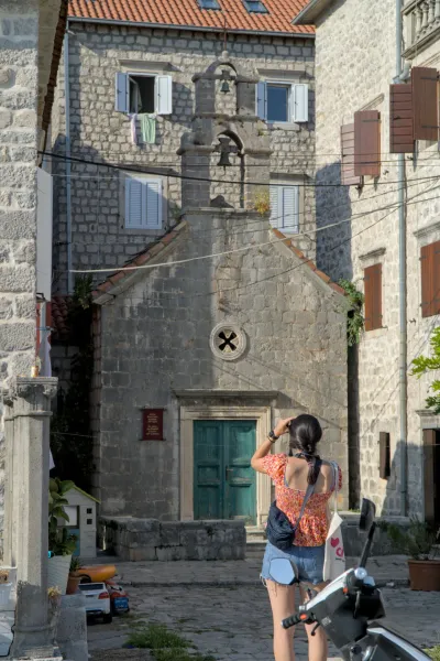 A woman photographs the "Alley of Perast" in Kotor, Montenegro, with a stone church facade and Venetian architecture.