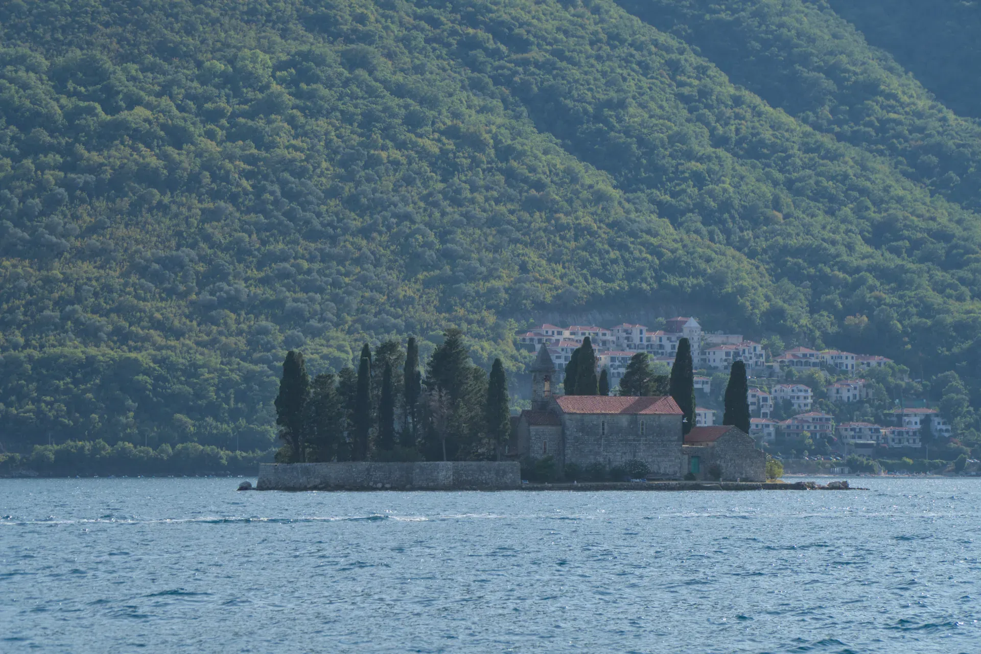 The St. George Monastery on an island in Perast, Montenegro, with lush greenery in the background and calm water in the foreground.
