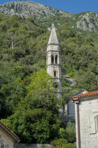 A stone bell tower rises from lush green vegetation, with rocky slopes in the background, on a clear evening in Perast.