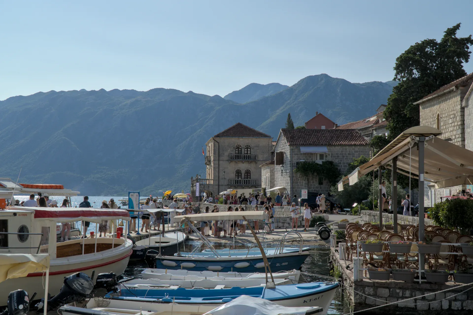 Un día soleado en el muelle de Perast, Montenegro, con barcos, edificios históricos y montañas al fondo.