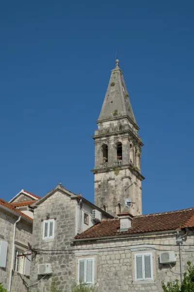 The stone bell tower of Perast stands against a clear blue sky, flanked by traditional houses.