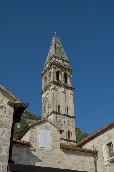 The famous bell tower of Perast, Montenegro, under a bright blue sky, with stone buildings in the foreground.