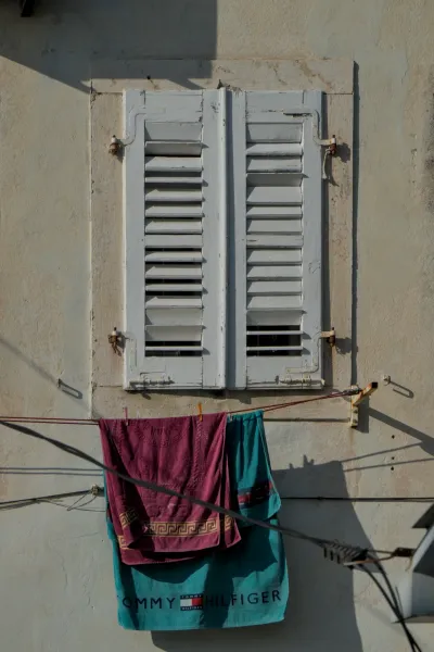 A window with white wooden shutters in Perast, Montenegro, with colorful laundry hanging on a line.