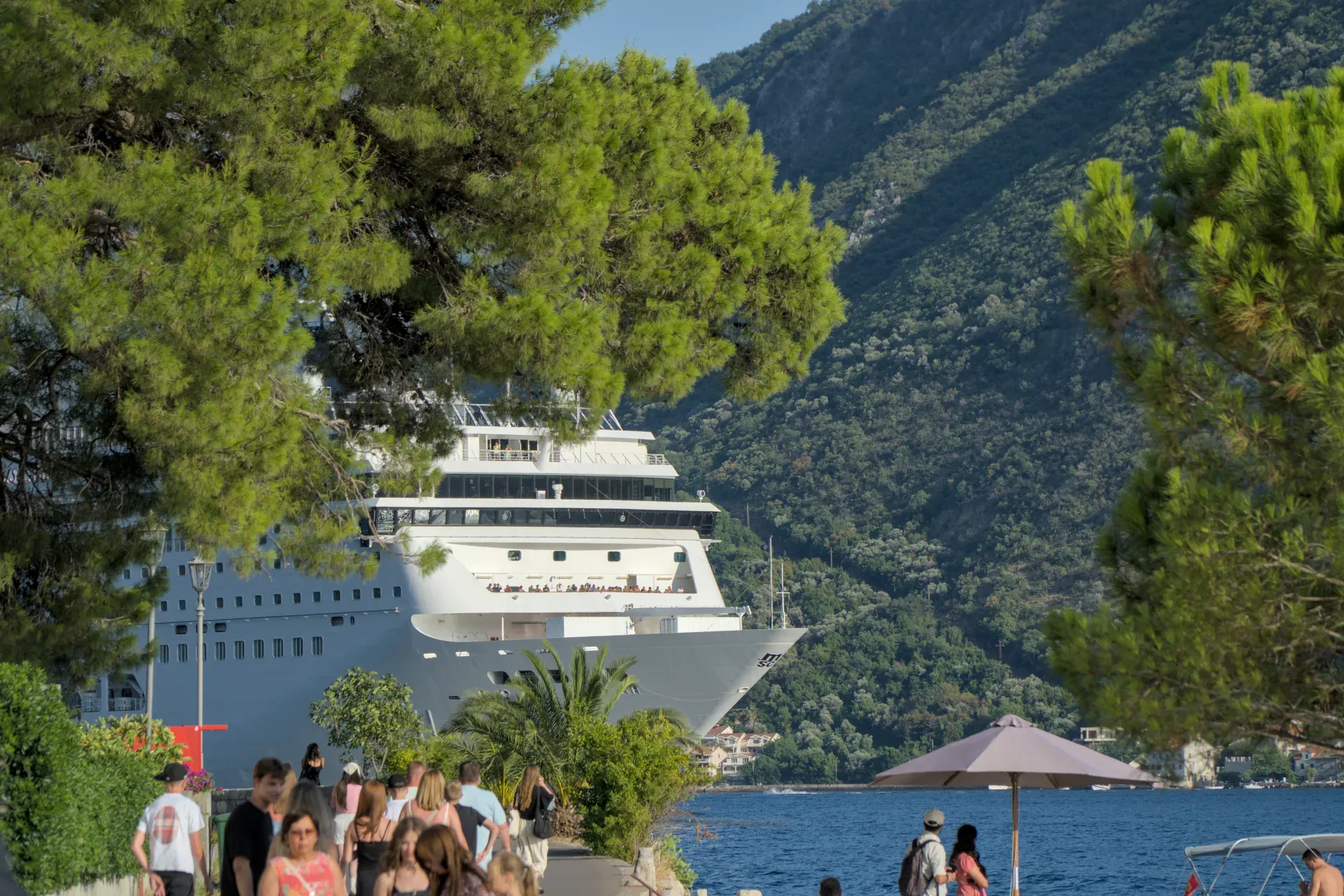 A giant cruise ship leaving the Bay of Kotor, with green trees and people on the shore in the foreground.