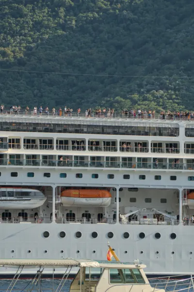 Passengers on the upper deck of a cruise ship wave goodbye to Kotor with a green mountain background.
