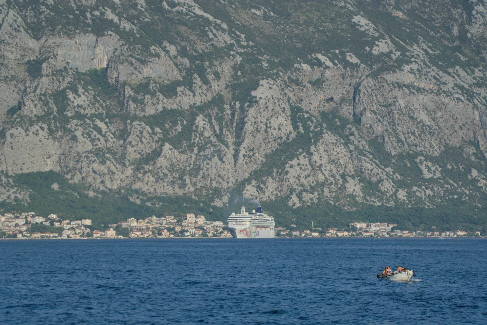 Un crucero y pequeños barcos en el mar frente a la ciudad de Kotor con sus casas y las montañas detrás.