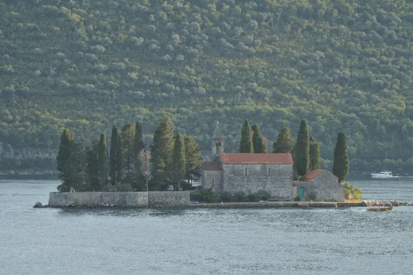 The St. George Monastery in Perast, Montenegro, on an island with cypress trees and calm water, against a forested hillside.
