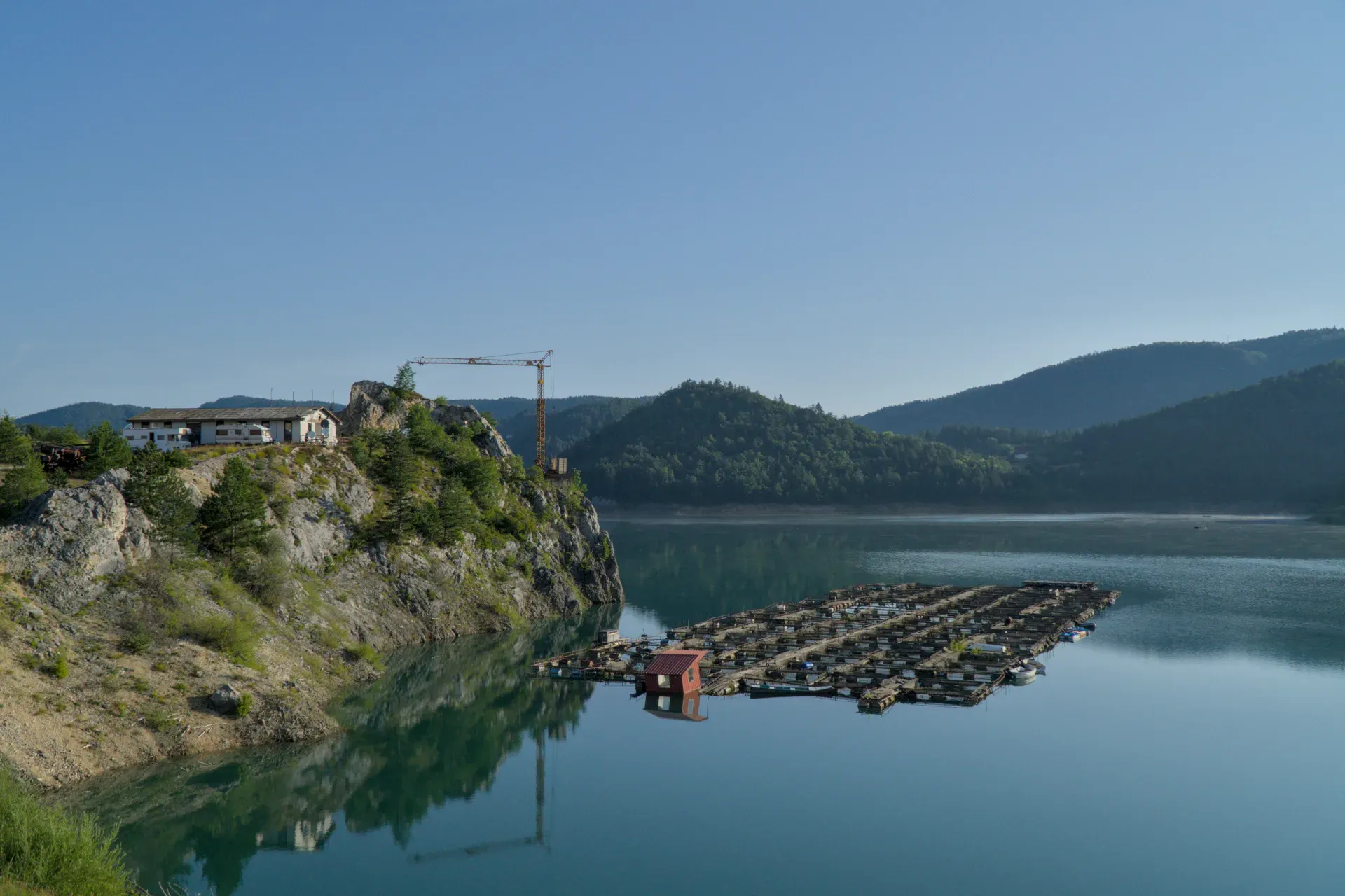 Sunrise at Zaovine Lake, Serbia. Calm water reflects mountains and clear sky with floating structures.