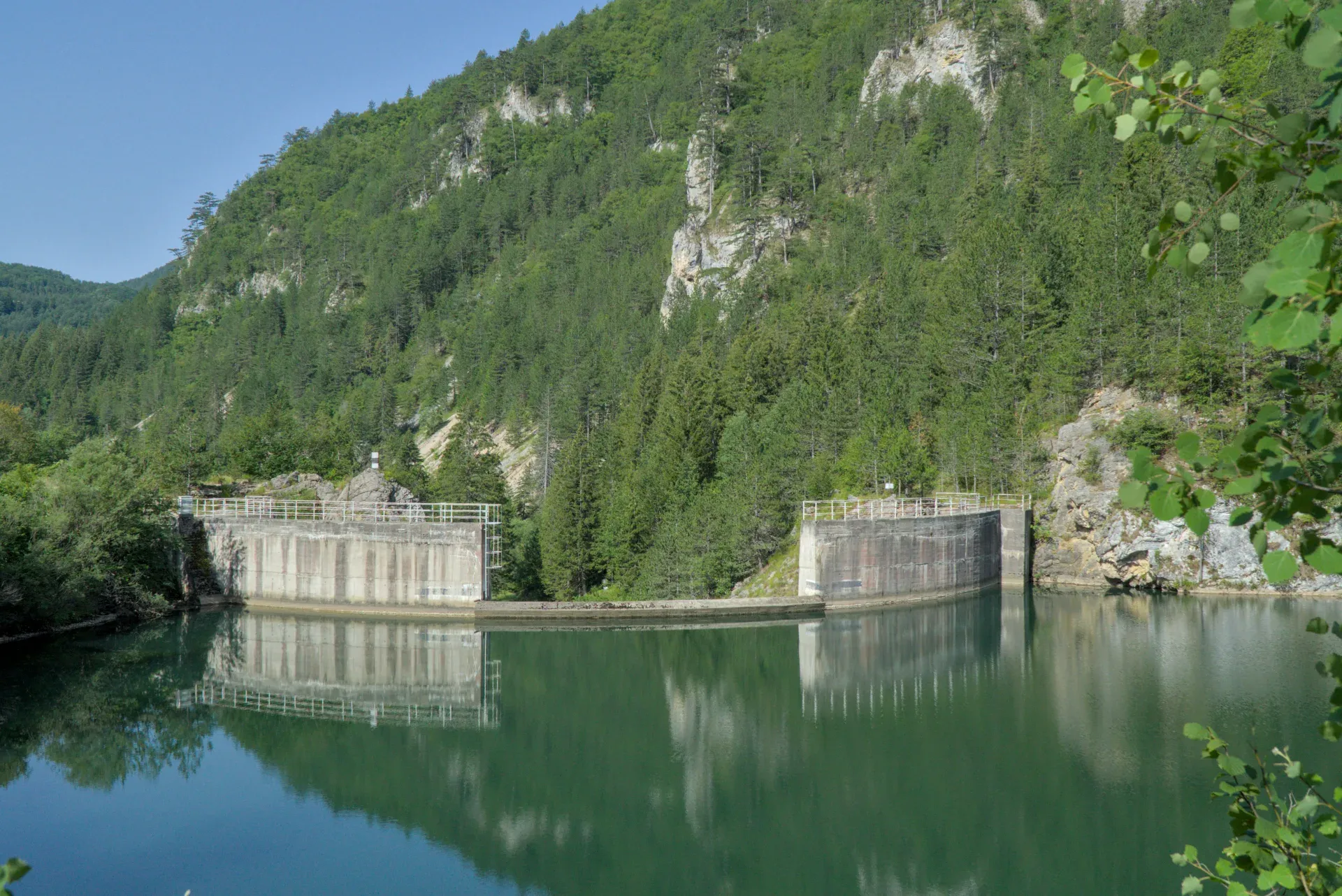 Le lac Spajići avec sa structure de barrage en béton, reflété dans des eaux vertes sur des collines boisées.