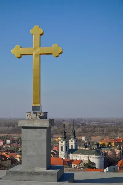 A prominent golden cross stands against a clear blue sky, overlooking a panoramic view of Sremski Karlovci, Serbia.