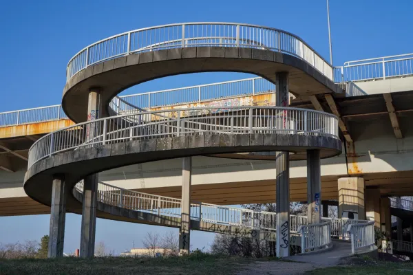 A concrete spiral ramp with metal railings, descending from the Liberty Bridge towards Kamenički Park.