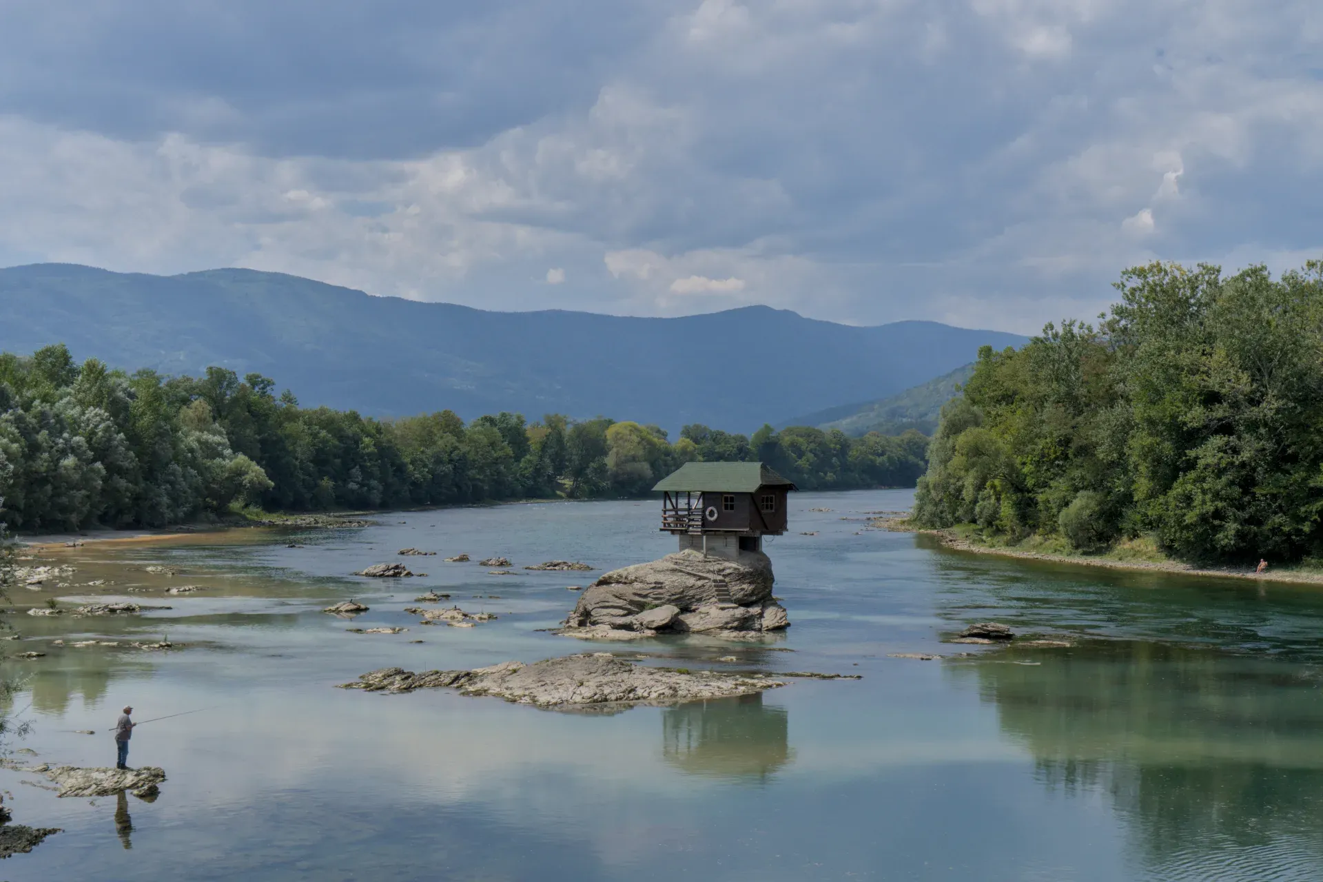 Une petite maison perchée sur un rocher dans la rivière Drina, avec un pêcheur solitaire lançant une ligne à proximité.