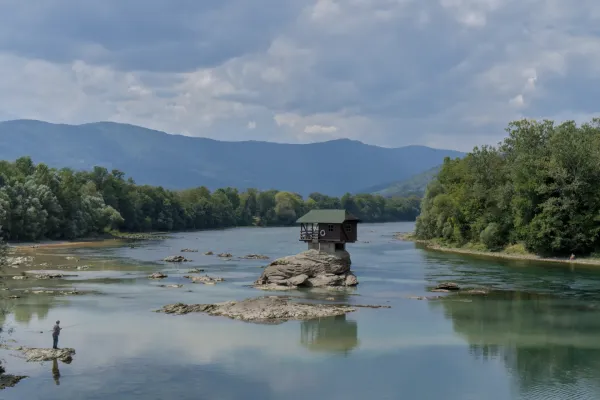 A small house perched on a rock in the Drina River, with a lone fisherman casting a line nearby.