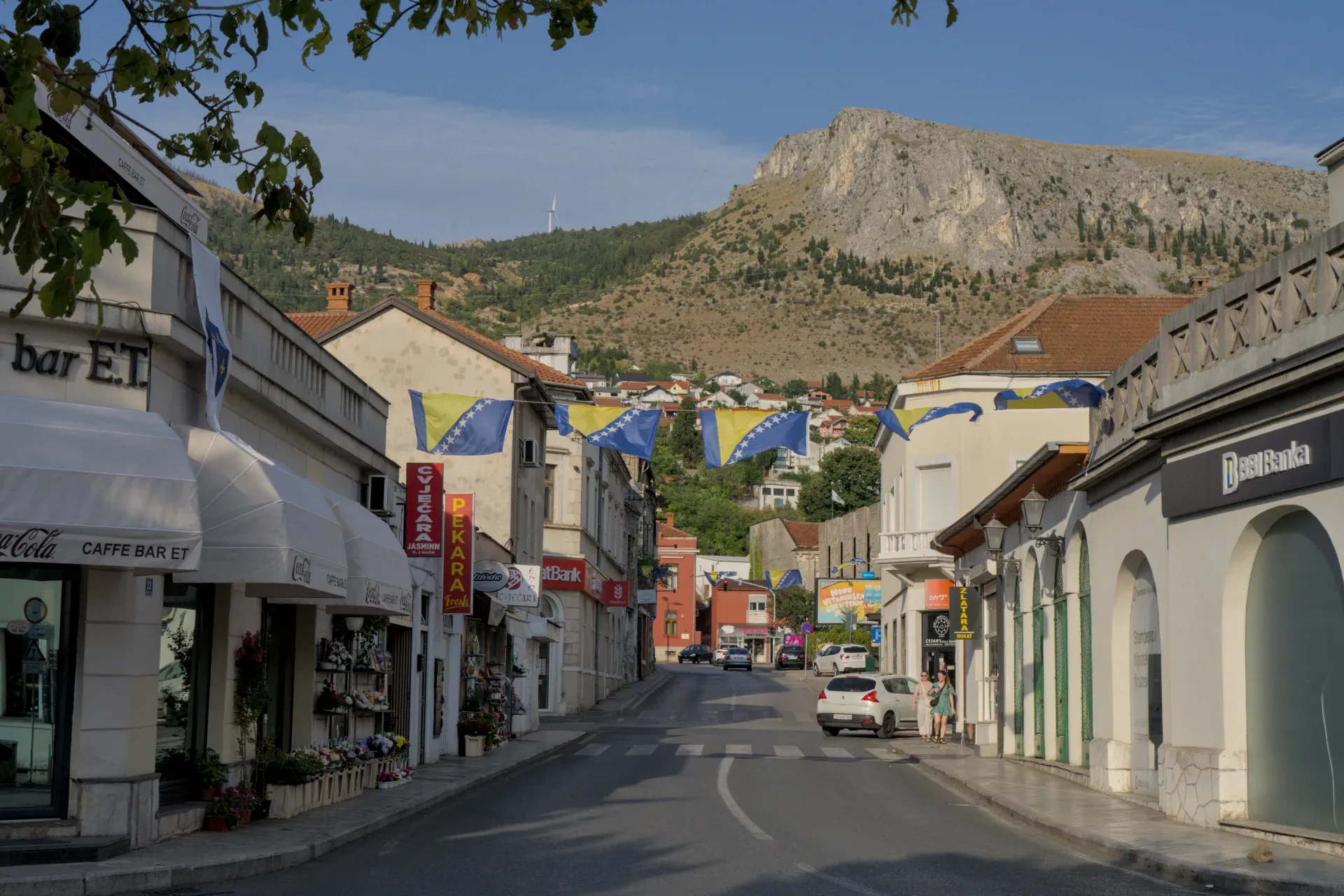 Street in Mostar with Bosnian flags before sunset, mountain backdrop.