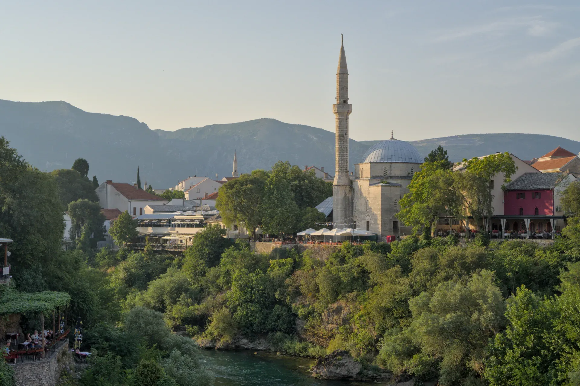 Fotografia paesaggistica dell'ora dorata della città vecchia di Mostar, con una moschea e vegetazione rigogliosa.