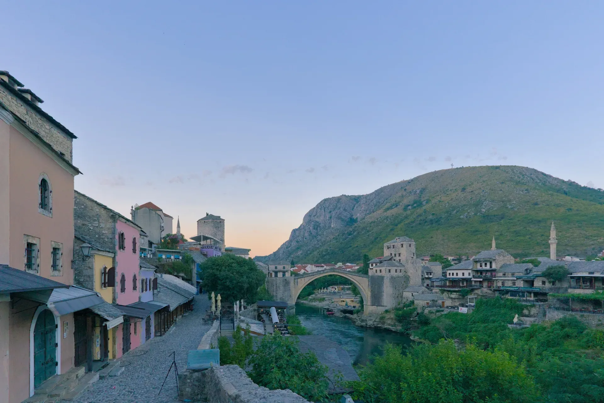 Panorama pre-alba della città vecchia di Mostar con case colorate, il ponte storico e un fiume calmo.