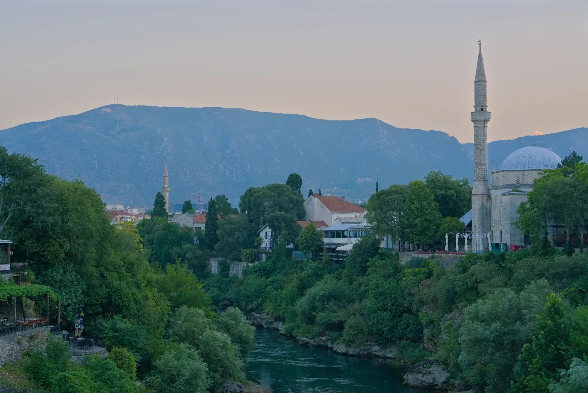 Casco antiguo de Mostar antes del amanecer, con el río Neretva, exuberante vegetación y una mezquita, capturado en formato horizontal.