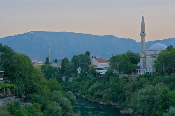 Mostar Old Town before sunrise, featuring the Neretva River, lush greenery, and a mosque, captured in landscape orientation.
