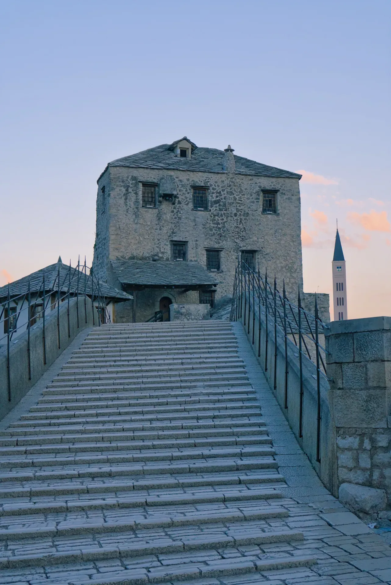 Stari Most u Mostaru u zoru, sa kamenim stepenicama i istorijskim zgradama u pozadini.