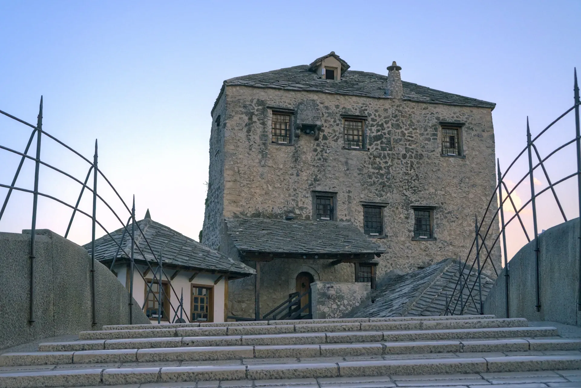 An ancient stone tower and adjacent buildings by the Old Bridge in Mostar, Bosnia and Herzegovina, with stone steps leading up.