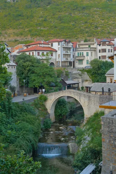 The "Crooked Bridge" in Mostar, Bosnia and Herzegovina, featuring a small waterfall and abundant greenery, in portrait orientation.