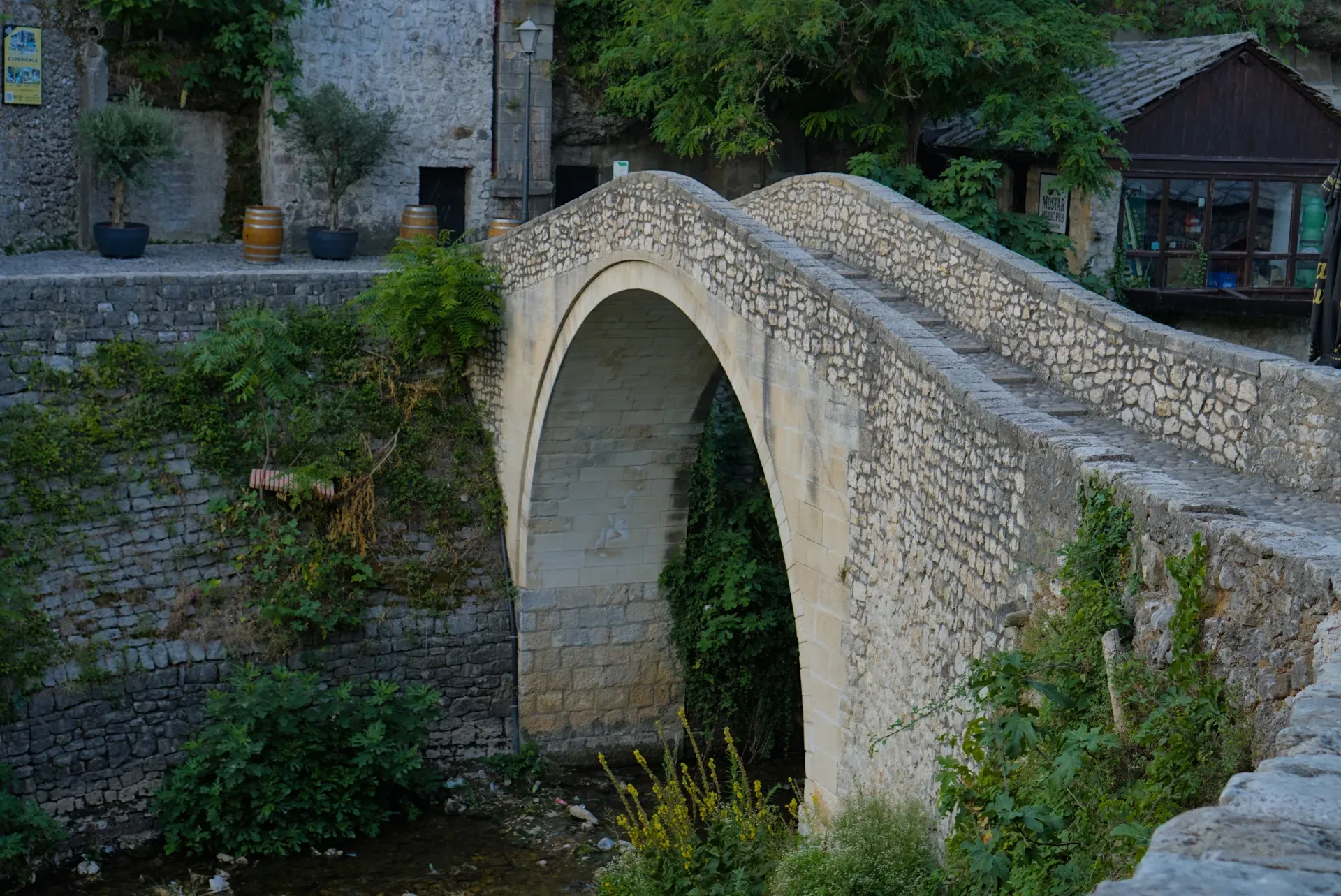 A stone bridge with a single arch, known as the 'krivi most' or crooked bridge, in Mostar, Bosnia and Herzegovina, surrounded by greenery.