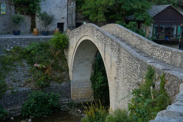 A stone bridge with a single arch, known as the 'krivi most' or crooked bridge, in Mostar, Bosnia and Herzegovina, surrounded by greenery.