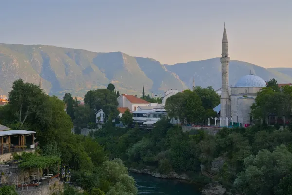 Landscape photograph of Mostar during golden hour, featuring a river, mosque, and mountains.