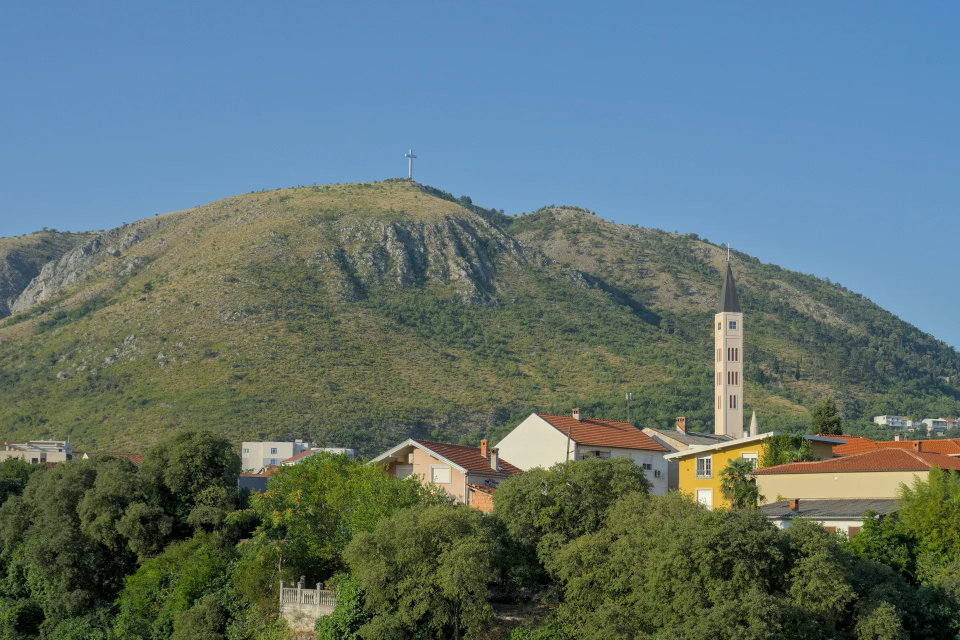 Panoramski likovni pejzaž Mostara sa velikim krstom na brdu i vitkim crkvenim tornjem u prvom planu.