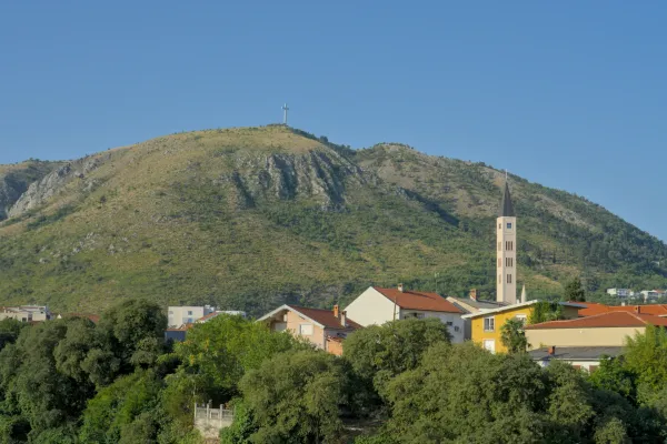 A panoramic fine art landscape of Mostar featuring a large cross on a hill and a slender church tower in the foreground.