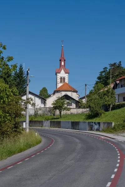 A winding road leads towards a church with a prominent red steeple in Predjama, Slovenia, under a clear blue sky.
