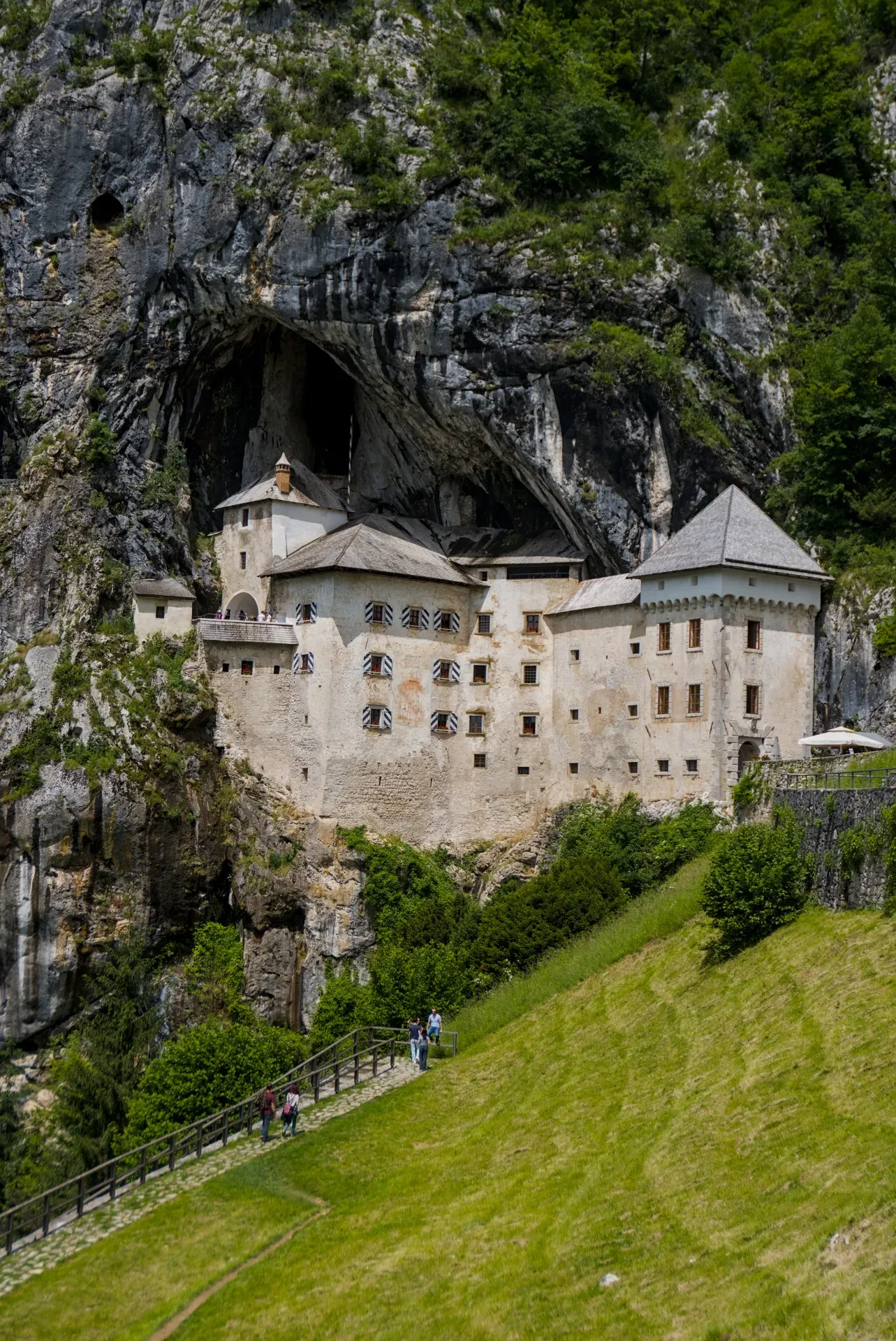 Predjama Castle nestled into a massive rock face, with green foliage and visitors on a path below.