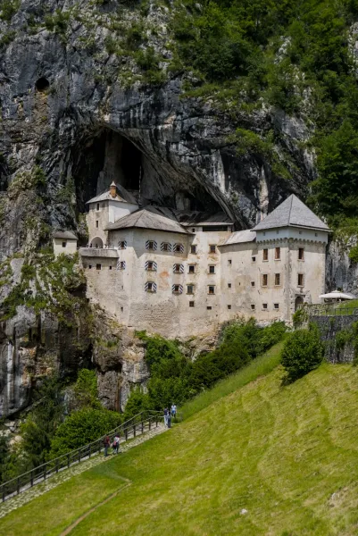 Predjama Castle nestled into a massive rock face, with green foliage and visitors on a path below.