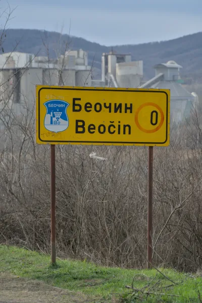Yellow road sign for 'Beočin' in Serbian and Latin script with a cement factory in the background.