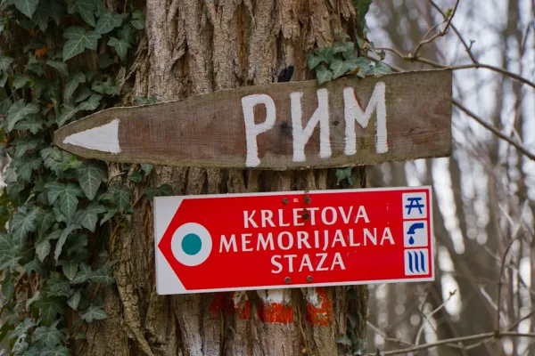 A wooden sign pointing left with Cyrillic text and a red sign for Krletova Memorijalna Staza on a tree in Fruska Gora.