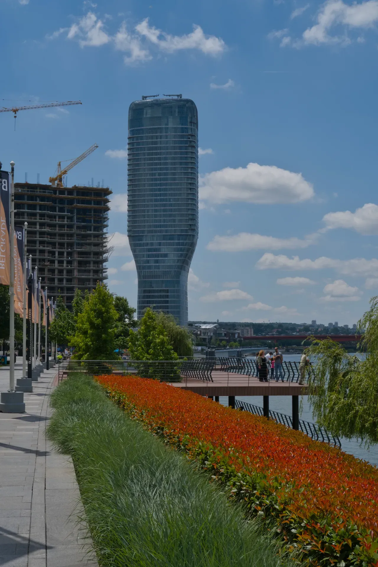 Modern building by the river with bright red flowerbeds and people on the promenade in Belgrade.