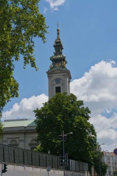 Elegant church bell tower in Belgrade against a blue sky with clouds, framed by trees.