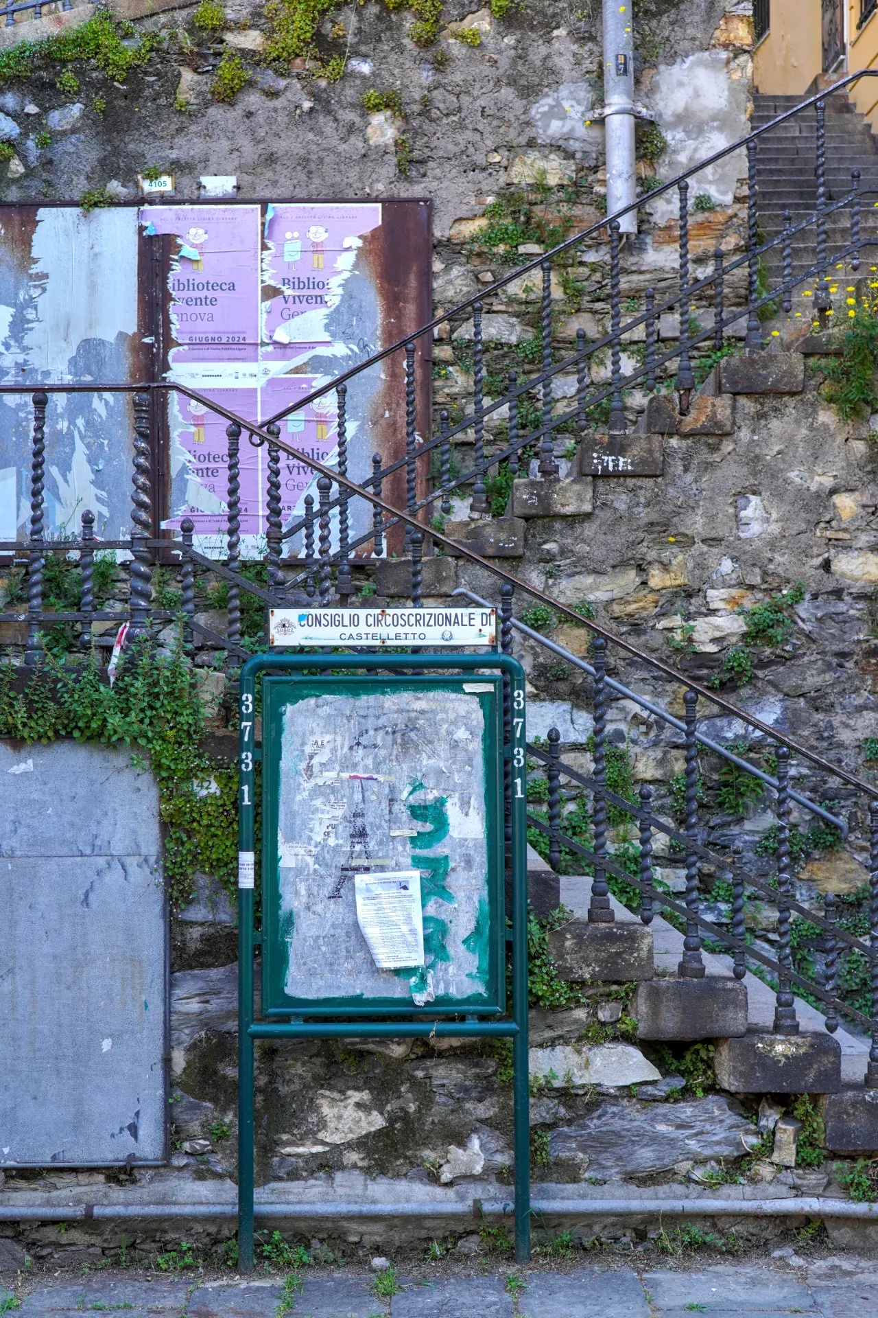 An old stone staircase with an ornate metal railing leads upwards next to a stone wall. In the foreground, a green bulletin board displays weathered posters, and overgrown plants climb the wall and stairs.
