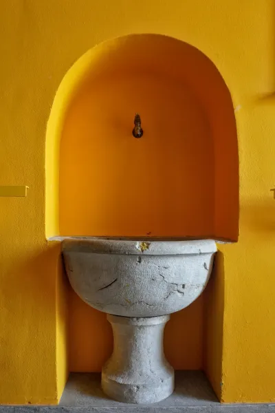 Un lavabo in pietra vintage si trova in una nicchia ad arco giallo brillante con un piccolo rubinetto in ottone sopra, contro una parete gialla testurizzata. Il pavimento è rivestito di piastrelle grigie tenui.