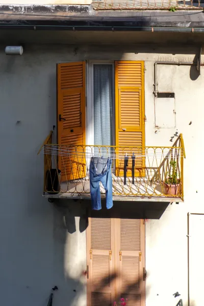 Ein Balkon in Genua, Italien, mit leuchtend orangen Fensterläden am Fenster, blauen Jeans und schwarzen Socken an einer Wäscheleine und einer kleinen grünen Topfpflanze.