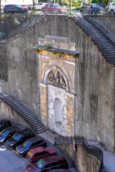 Ein verwittertes historisches Denkmal mit einer skulpturalen Reliefdarstellung unter einem Bogen, vor einer rauen Steinmauer, mit zahlreichen Außentreppen und einem Parkplatz mit Autos darunter in Genua, Italien.