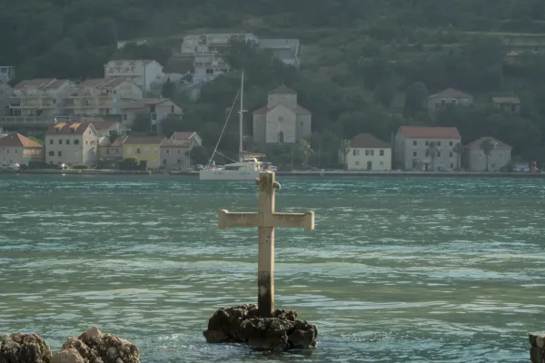 A stone cross on a rock in the water of the Bay of Kotor, Montenegro, with a coastal town and green hills in the background.