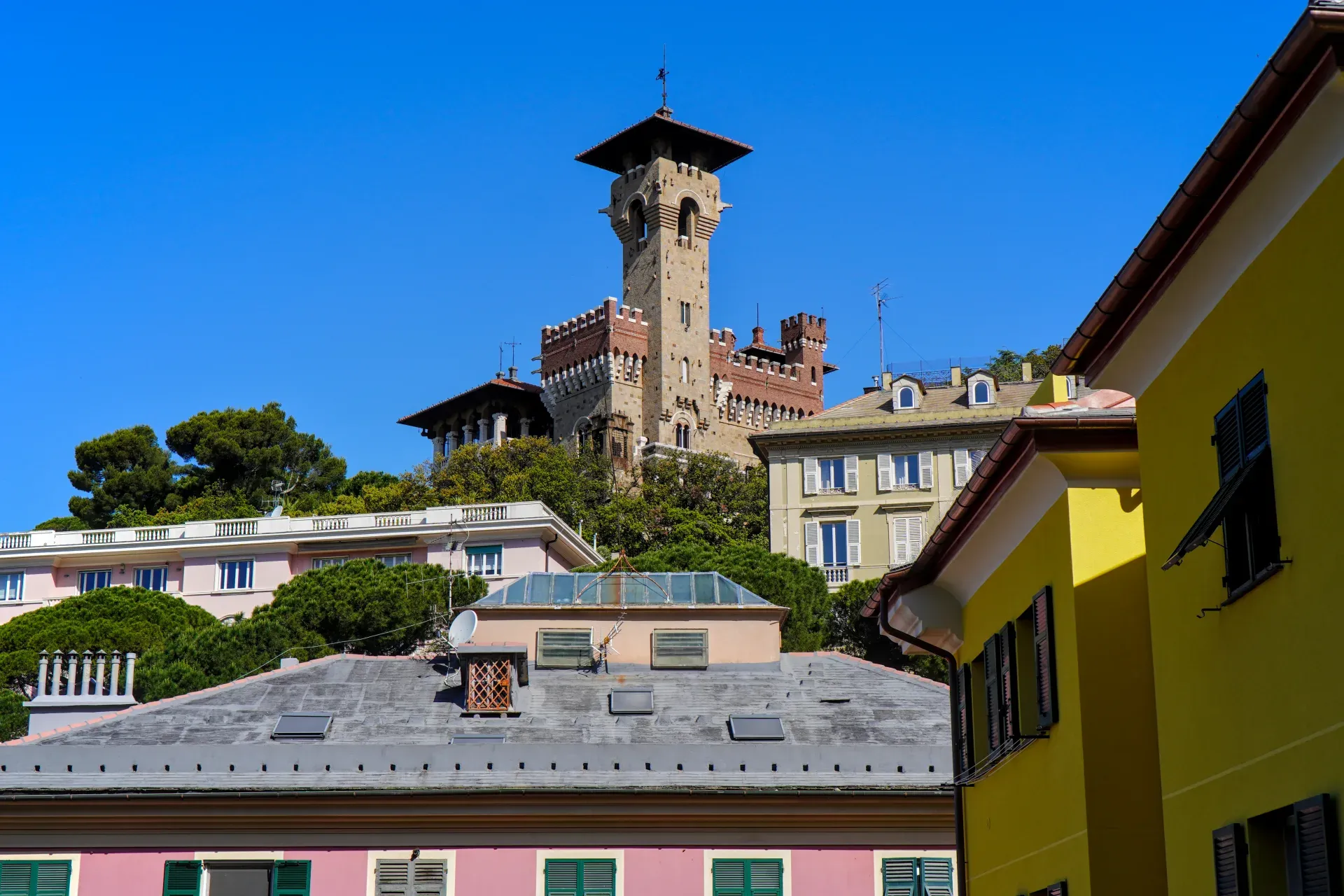 A view of Castello Mackenzie, a castle in Genoa, Italy, with its striking tower, set against a backdrop of green trees and residential buildings under a bright blue sky.