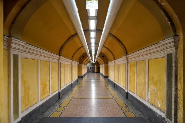 Ein langer, symmetrischer Tunnel mit gelben Wänden, weißen Zierleisten, einem gemusterten Marmorboden und heller Deckenbeleuchtung.