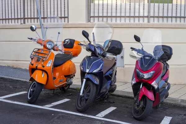 Ein oranger Vespa Roller, ein dunkelblauer Roller und ein roter Roller sind in einer Reihe auf einer Straße mit weißen Parklinien geparkt.