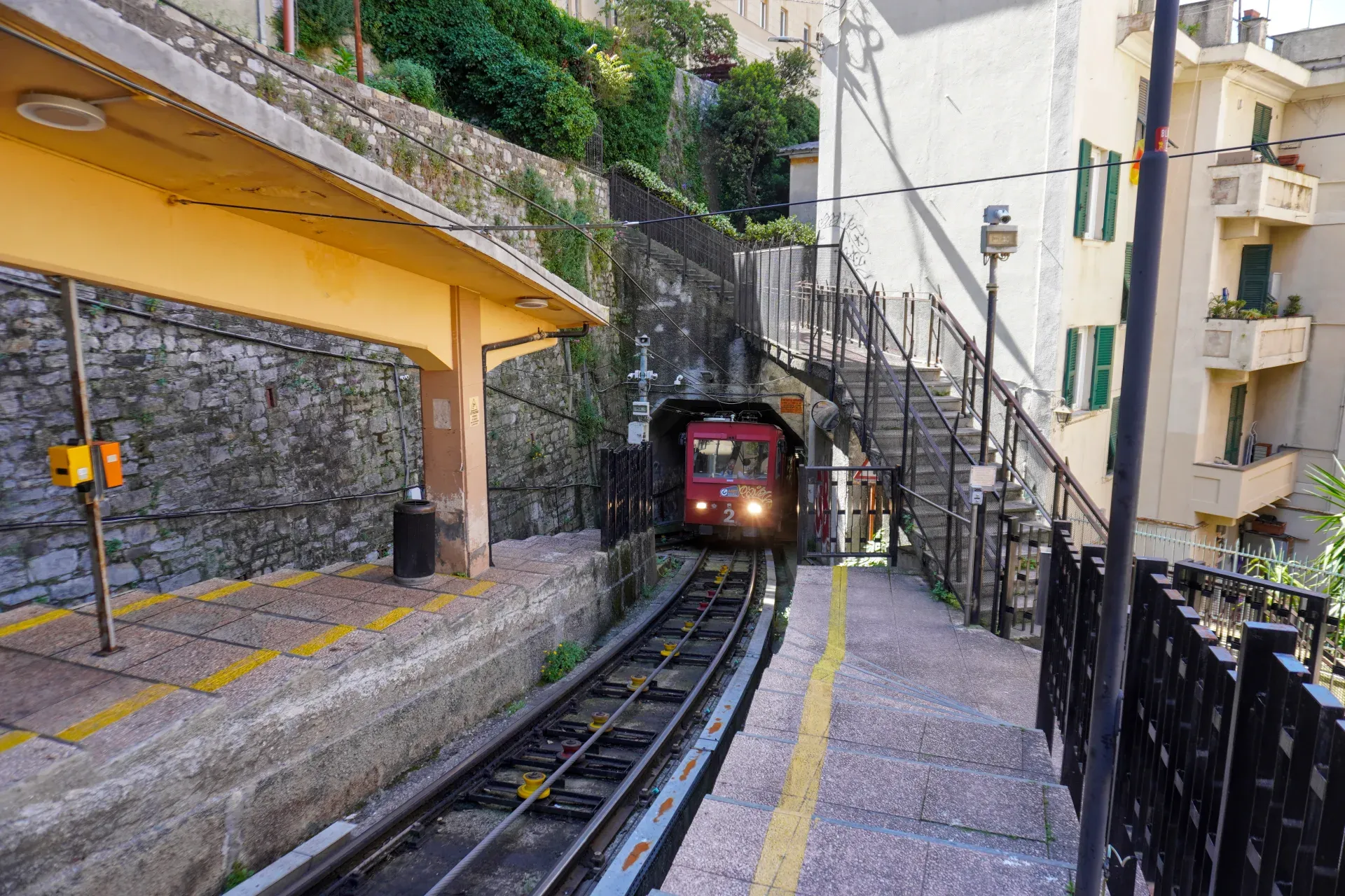 A red funicular with headlights on enters a tunnel on a steep railway track. The view is from the platform of a station with a yellow roof built against a stone wall. To the right, a staircase with a black railing leads up the slope next to a building with balconies.