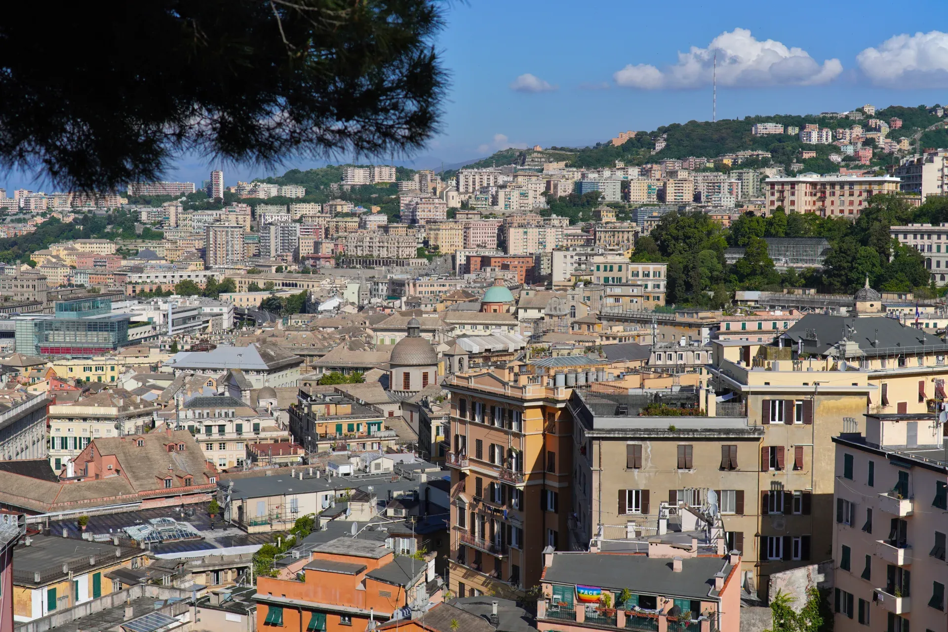 Pogled na šarene zgrade i zeleno drveće na brdima Đenove, Italija, sa istorijskim tornjem u pozadini pod vedrim plavim nebom.