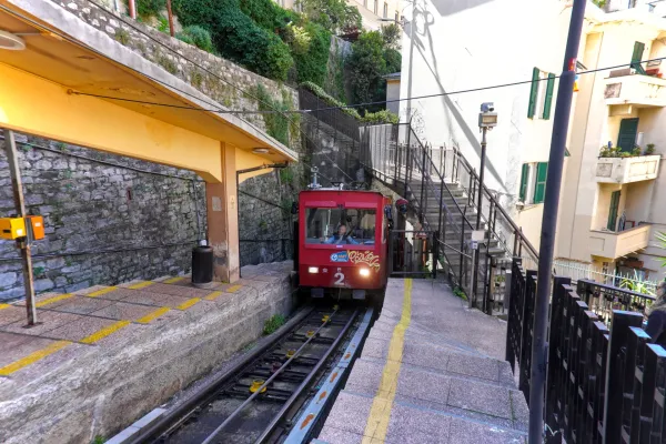 Ein roter Standseilbahnzug mit der Nummer 2 auf der Vorderseite fährt in Genua, Italien, auf einer steilen Strecke bergauf, neben einem Bahnsteig mit gelbem Dach.