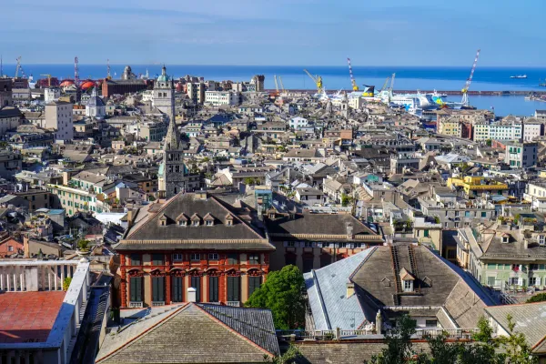 Una ripresa grandangolare di Genova, Italia, che mostra il porto attivo con navi cargo, traghetti e gru, affacciata sul denso paesaggio urbano che si estende verso il mare blu.